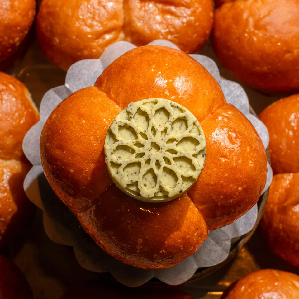 A brioche-style bun marked with a pressed pattern of herbed butter on its crown, resting in paper among a tray of golden buns.