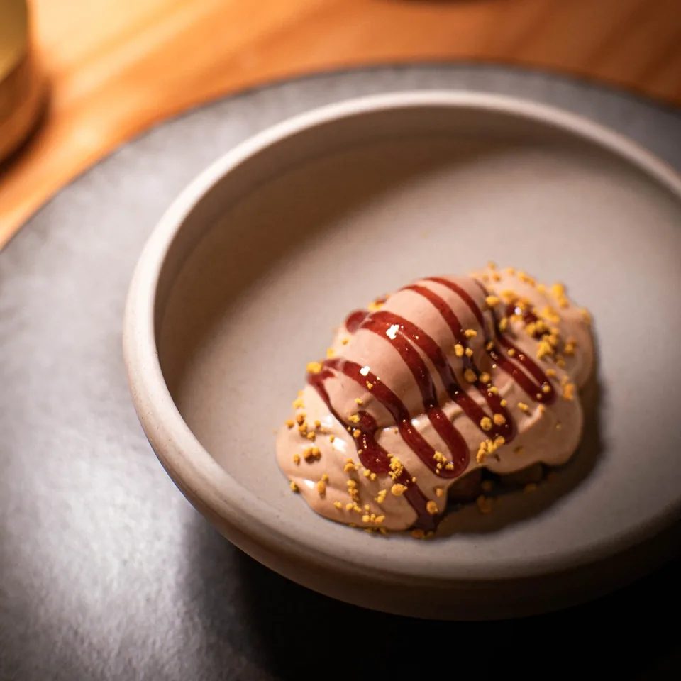 A swirl of pale chocolate cream striped with dark red syrup and flecks of gold crumb, in a grey stoneware bowl on a wooden table.